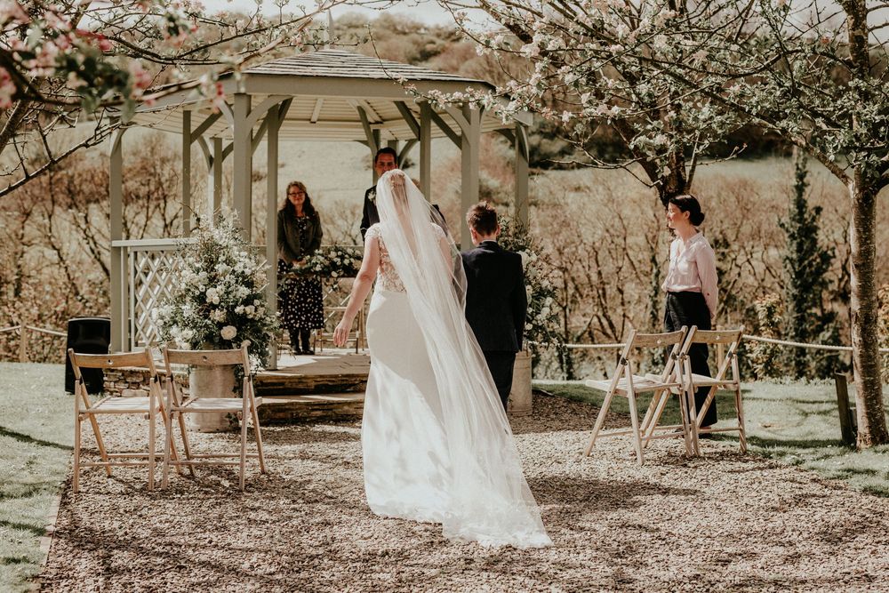 A rustic outdoor wedding ceremony under a wooden pergola with cherry blossoms and white chairs at golden hour.