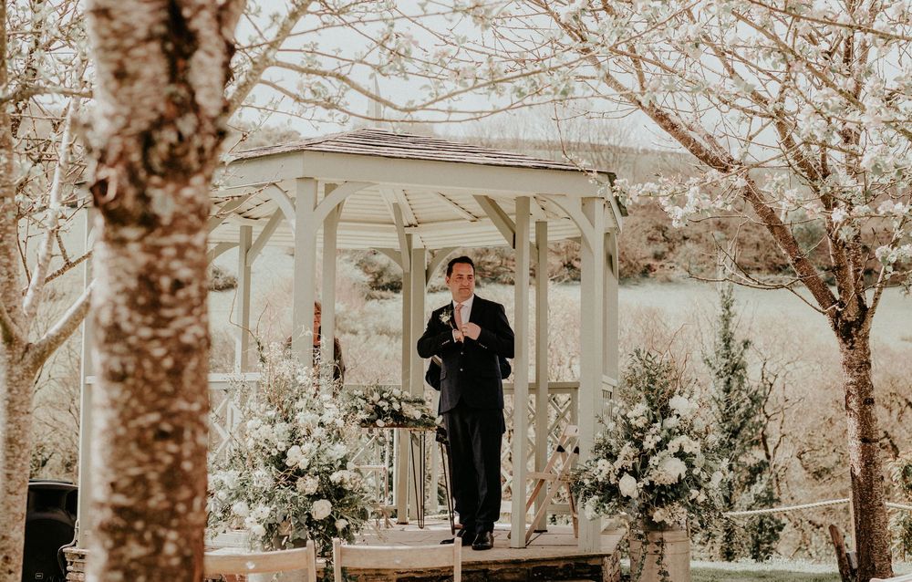 A figure in a tuxedo stands at a white gazebo decorated with floral arrangements for an outdoor wedding ceremony.