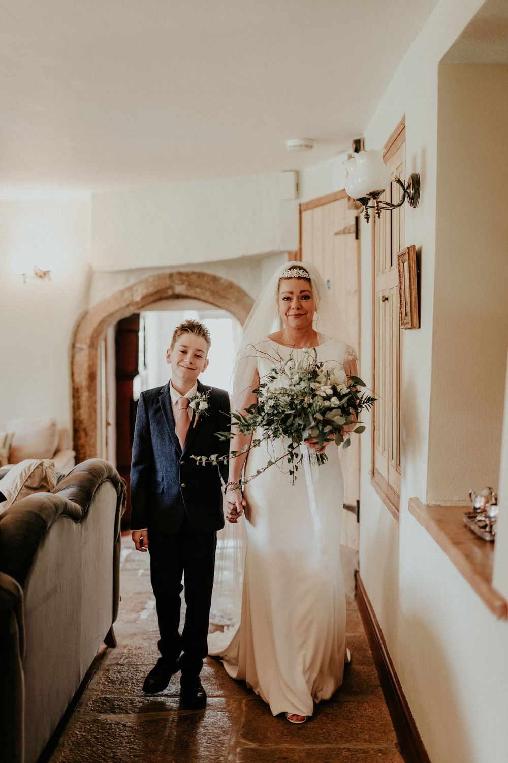 A wedding photo in a cozy hallway shows the bride in white dress holding a beautiful bouquet beside a young boy in suit.