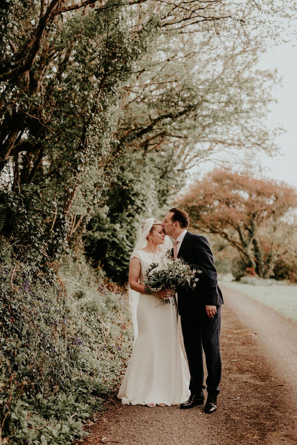 A couple shares a romantic moment on a tree-lined dirt path while holding a rustic floral bouquet on their wedding day.