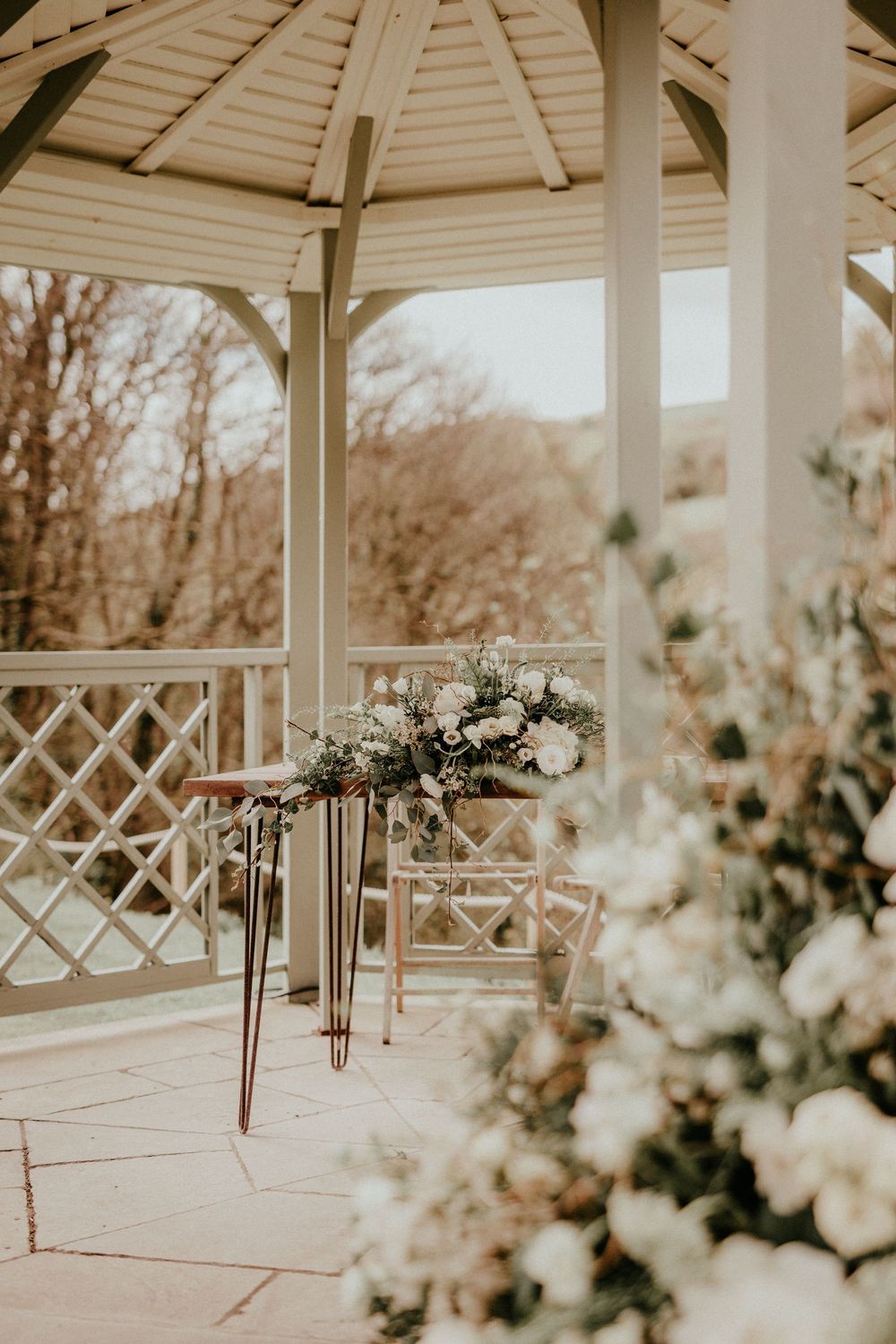 A white wooden gazebo decorated with floral arrangements and a small table for an intimate outdoor wedding setup.