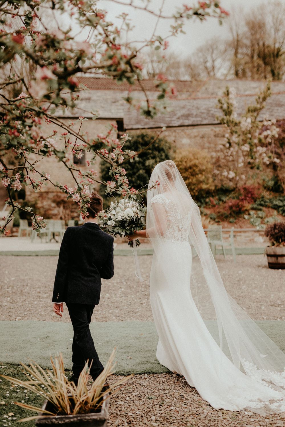 A couple walks through a garden path during an autumn wedding with blooming trees and a long white veil trailing behind.
