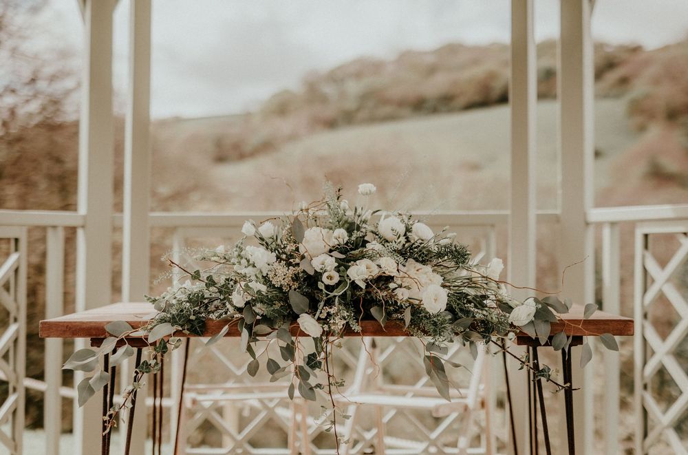 Elegant white floral wedding arch arrangement with greenery against a soft neutral backdrop at an outdoor venue.