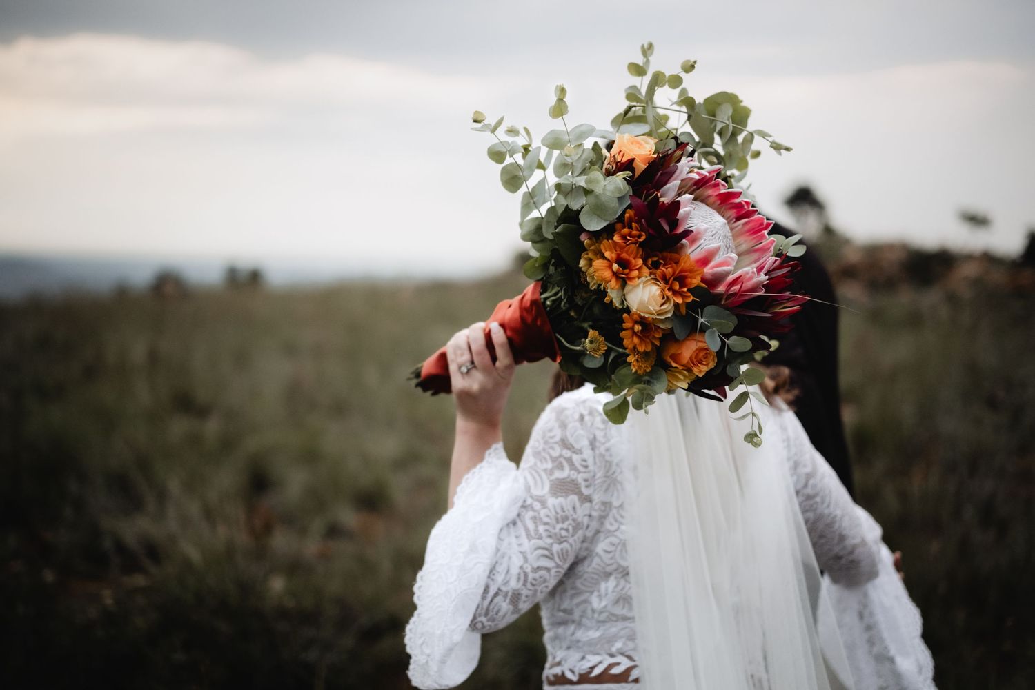Figure in a white lace dress holds a colorful autumn wedding bouquet with orange and red flowers outdoors.
