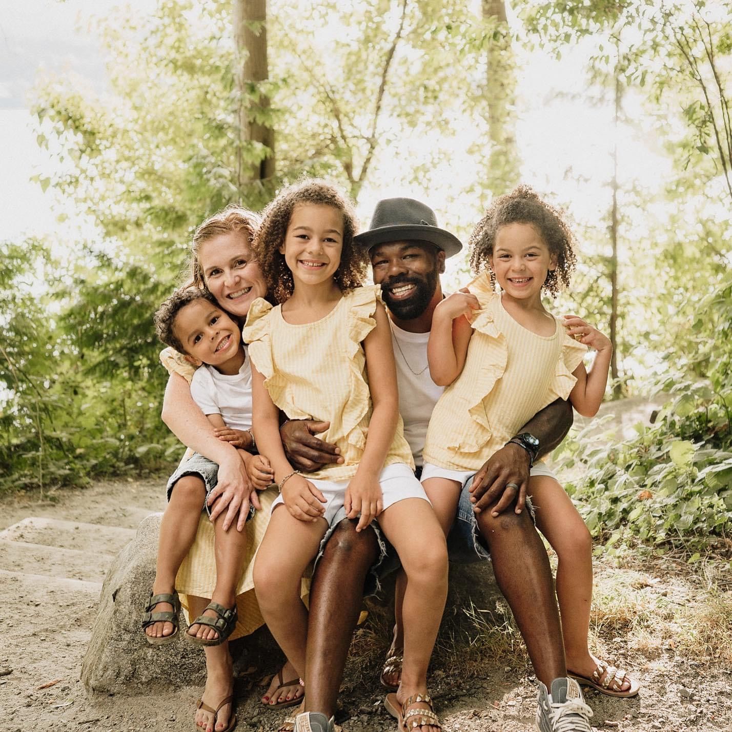 Family sitting together outdoors on a sunny day in a natural forest setting wearing casual summer clothing.