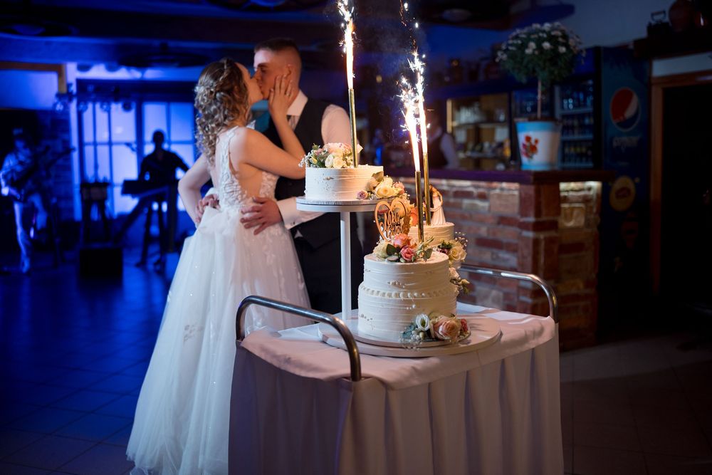 A wedding couple stands behind their sparkling wedding cake in a dimly lit reception venue with blue lighting.