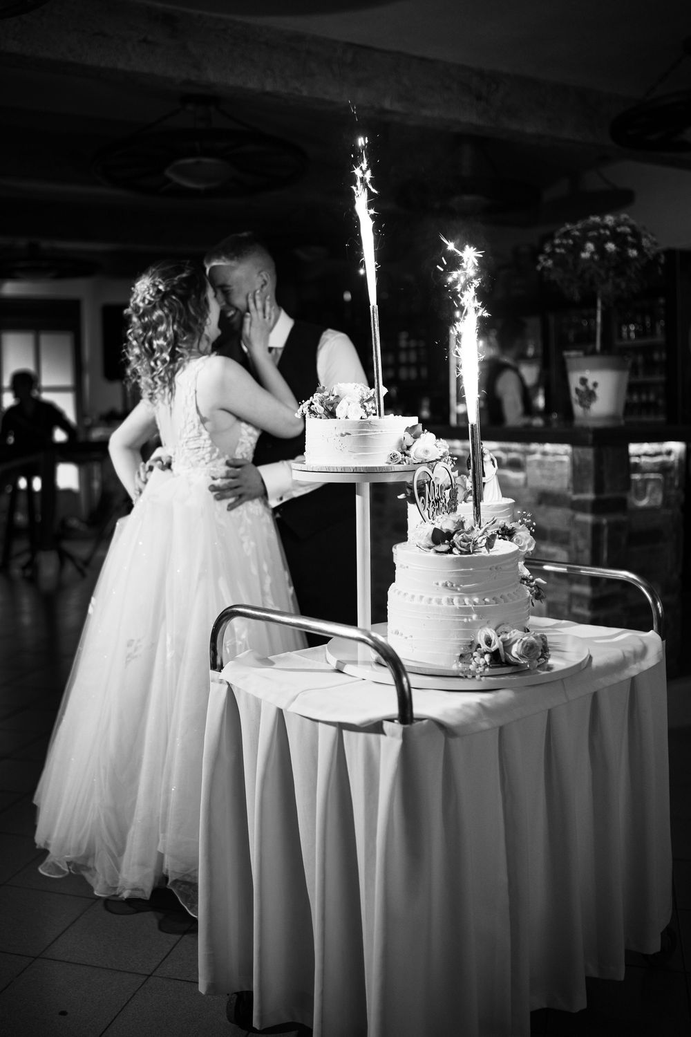 Black and white photo of a wedding reception showing a tiered cake with sparklers and couple embracing beside it.