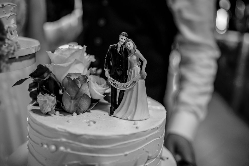 Black and white photo of a traditional wedding cake topper with bride and groom figurines and roses on top of a white cake.