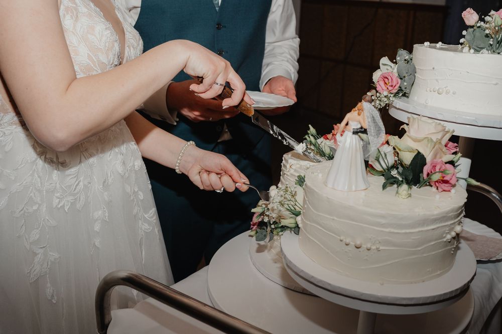 Two pairs of hands slice into a white tiered wedding cake decorated with pink roses and floral accents.