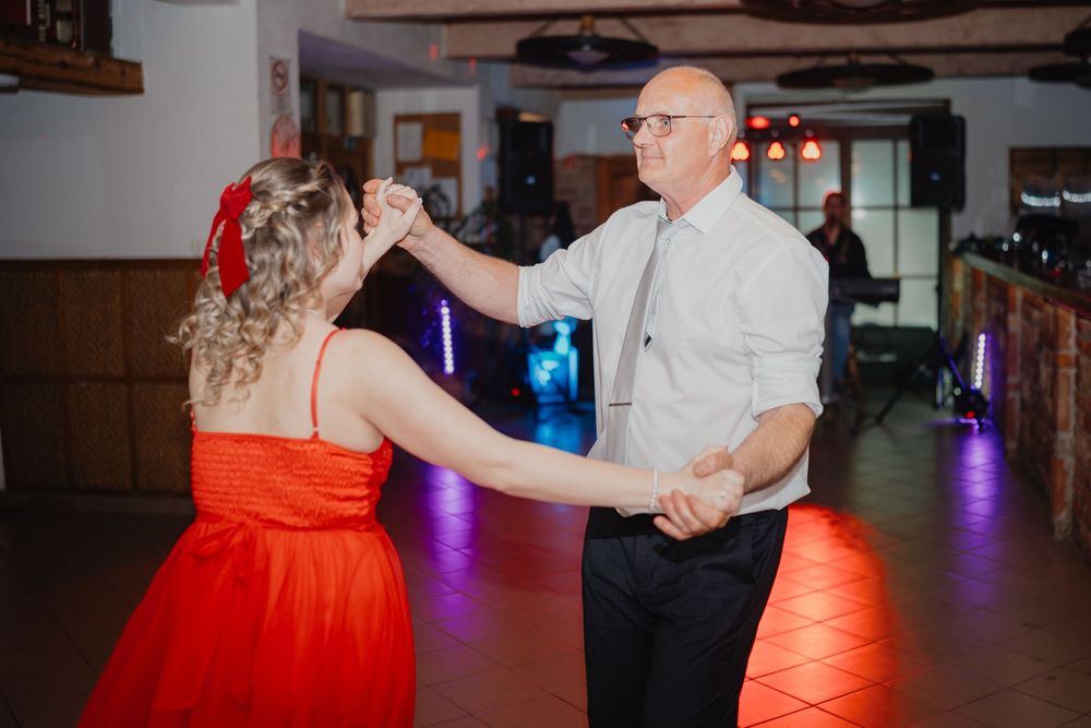 A couple dances together at an event, with the woman wearing a bright red dress and the man in a white button-down shirt.