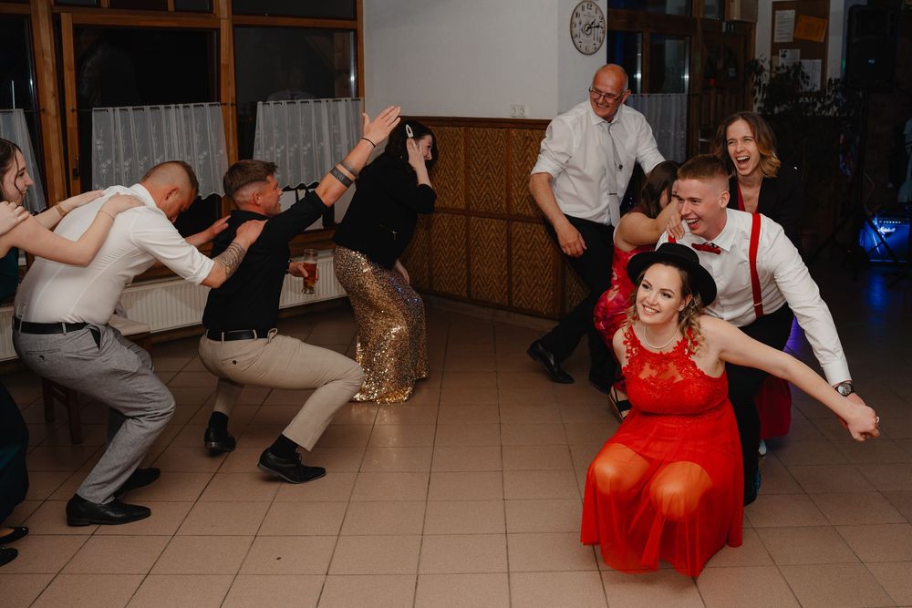 Party guests in formal wear dance energetically at a reception with tiled floors and wood paneled walls.