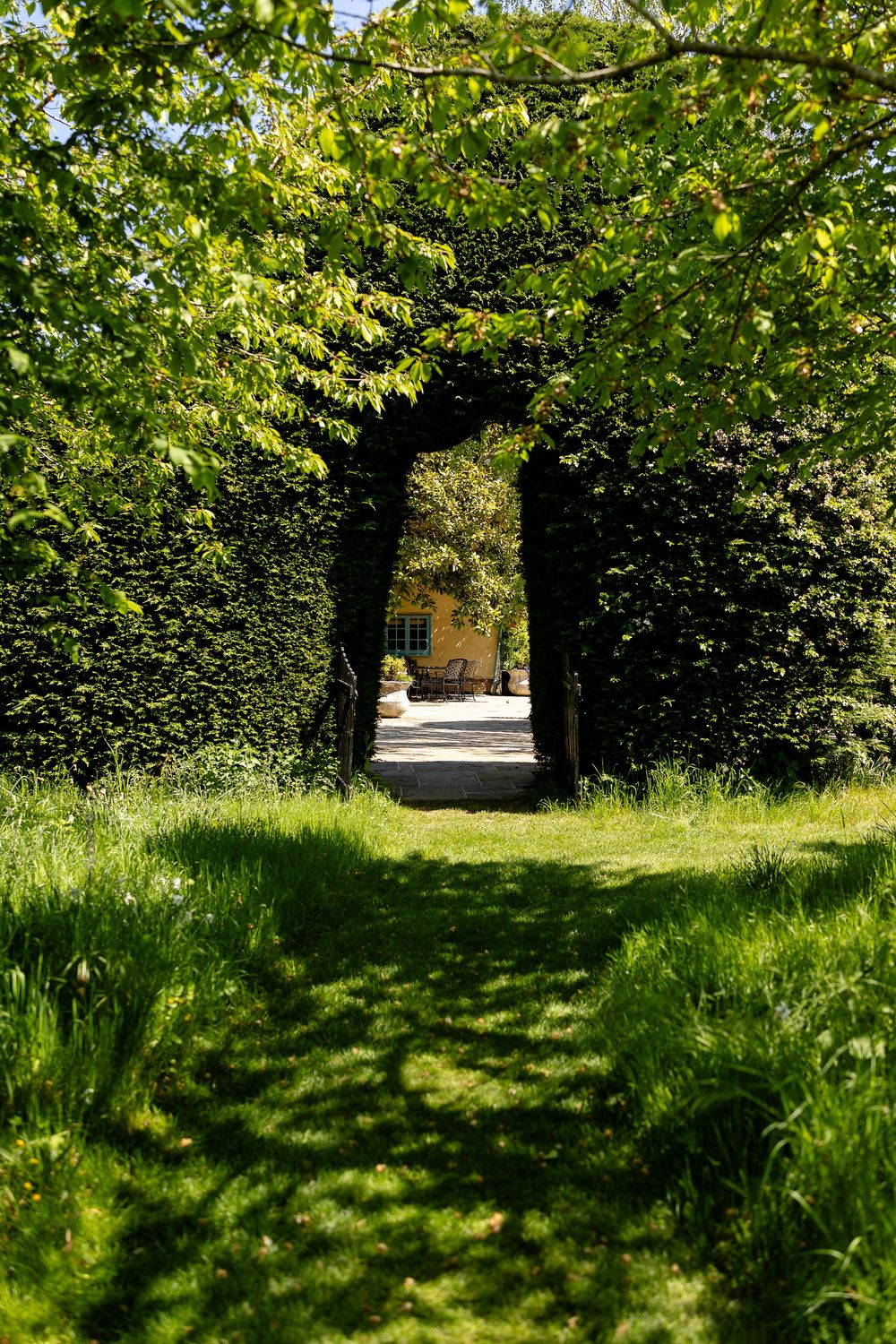A green garden archway formed by meticulously trimmed hedges creates a natural tunnel leading to a sunlit path.