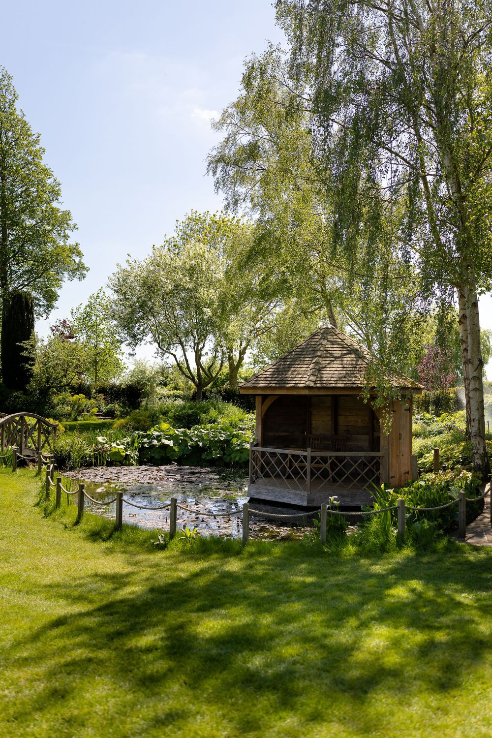 A rustic wooden gazebo sits beside a tranquil pond surrounded by lush green gardens and weeping willow trees.