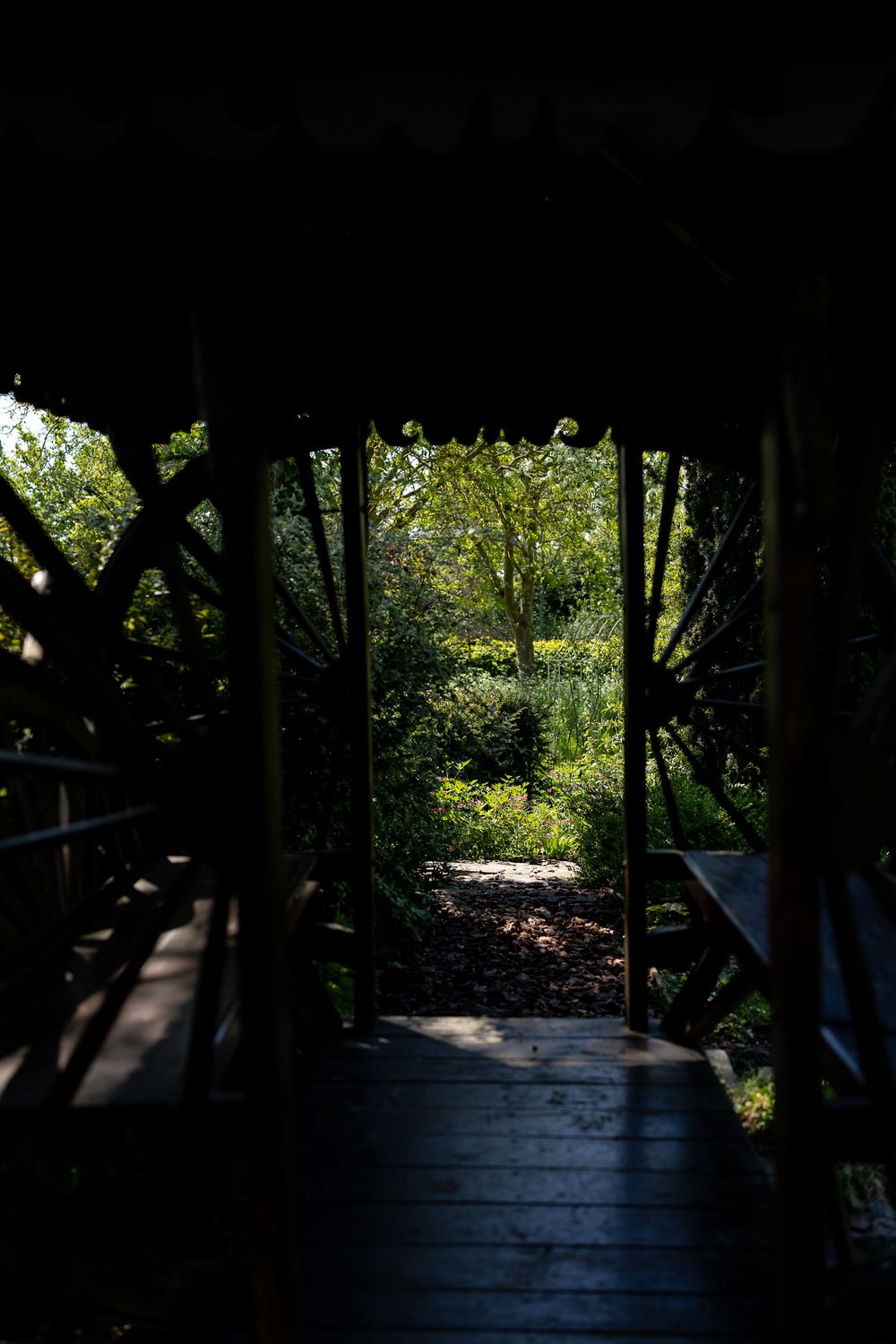 A wooden walkway through a dark tunnel opens to a sunlit garden path beyond.