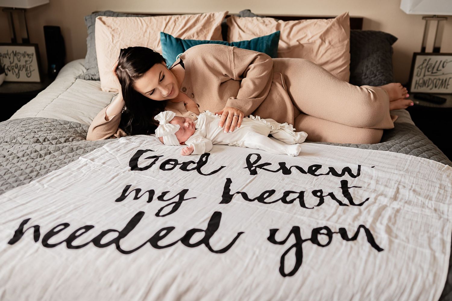 Mother lying on bed with text "God knew my heart needed you" written on blanket as she snuggles her newborn during in home newborn photos.
