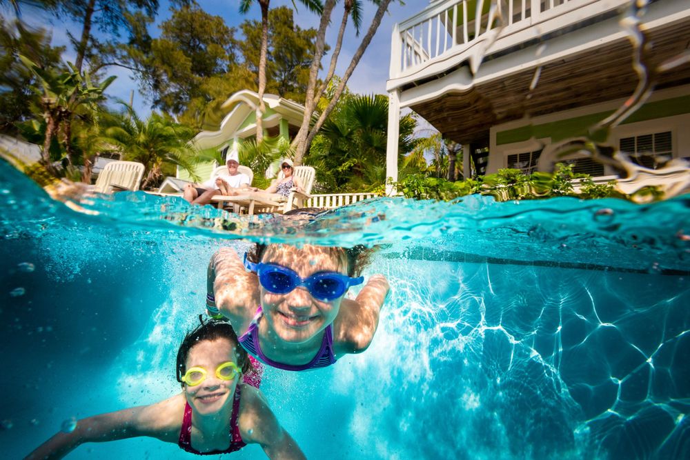 Underwater view of swimmers enjoying a pool on a sunny day with a house and tropical plants visible above.
