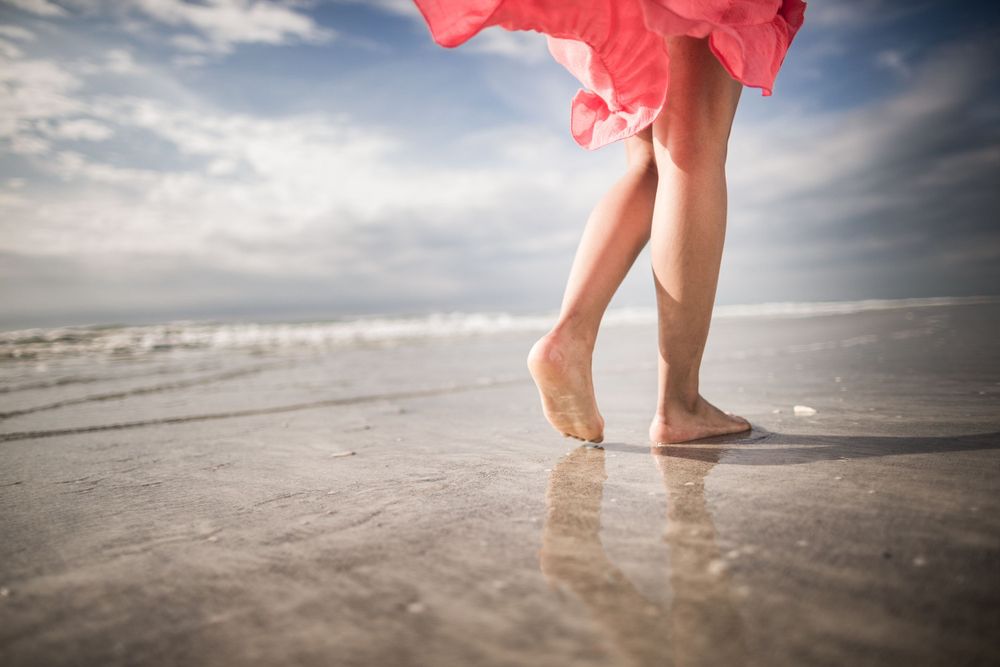 Bare feet in wet sand with flowing red dress on a beach at low tide.