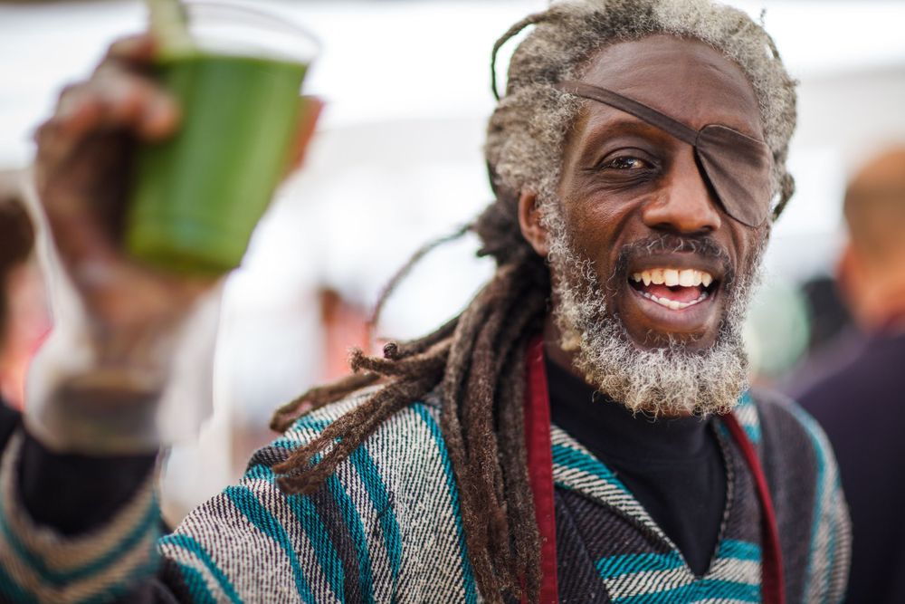 A joyful person with long dreadlocks and a gray beard holds up a green cup while wearing a striped sweater.