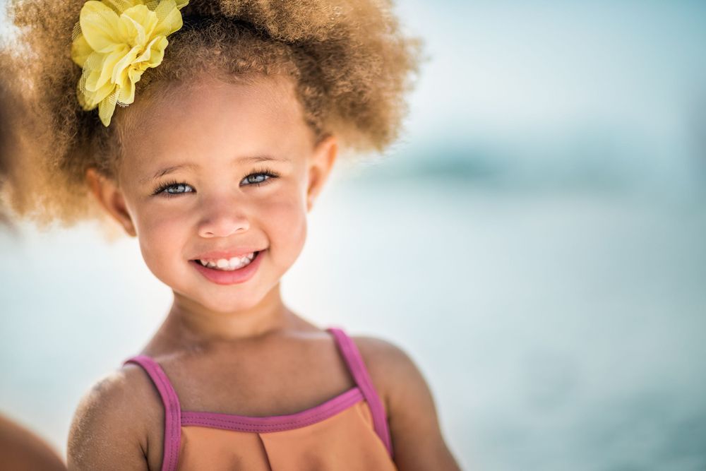 A joyful young child with curly hair and a yellow flower accessory smiling brightly.