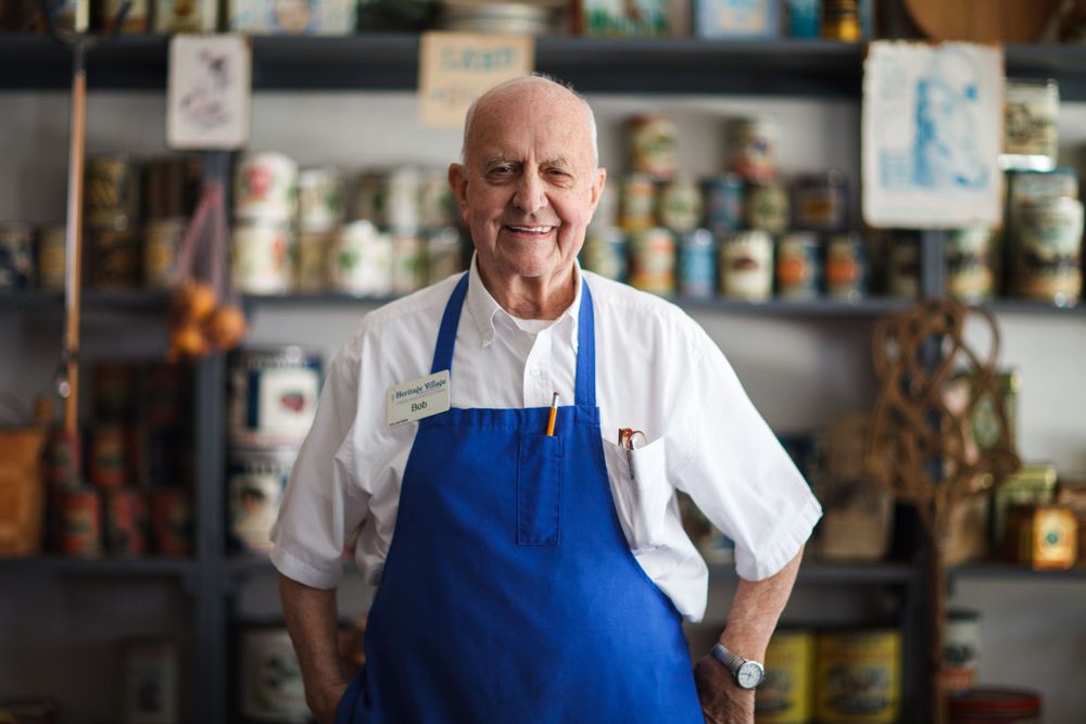 A smiling shopkeeper in a white chef coat and blue apron stands in front of shelves stocked with various goods and supplies.