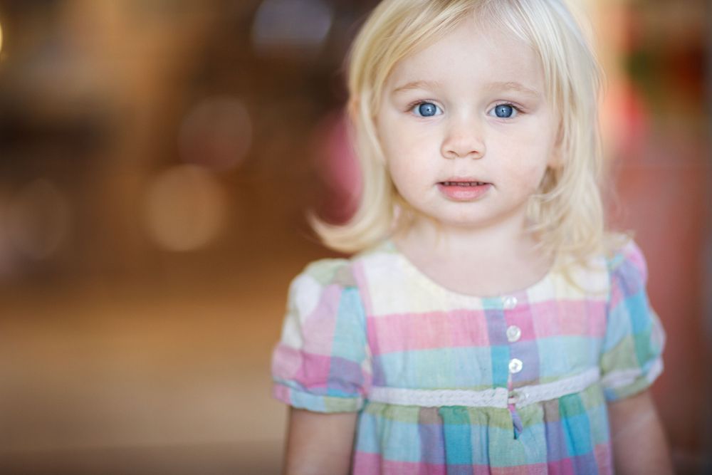 A young child in a pastel plaid dress looks at the camera against a soft blurred background.