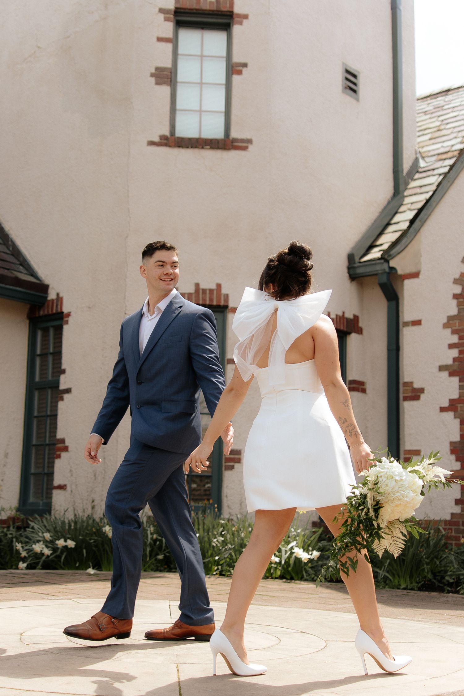 A couple in wedding attire walking together outside a stucco building with a white floral bouquet.