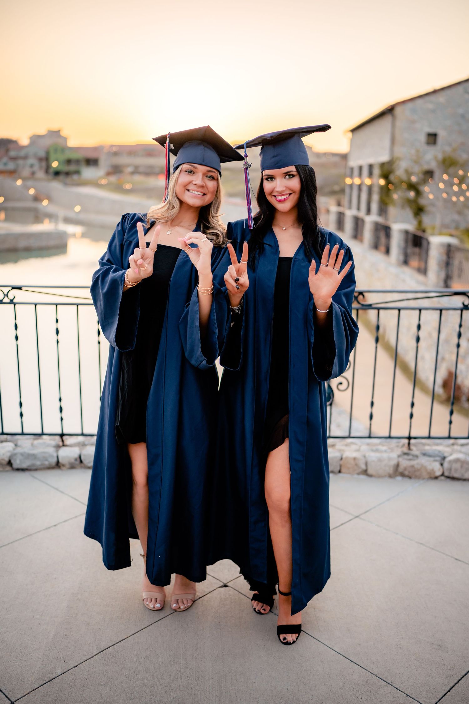 Two graduates in navy blue caps and gowns pose together on a balcony at sunset showing peace signs.