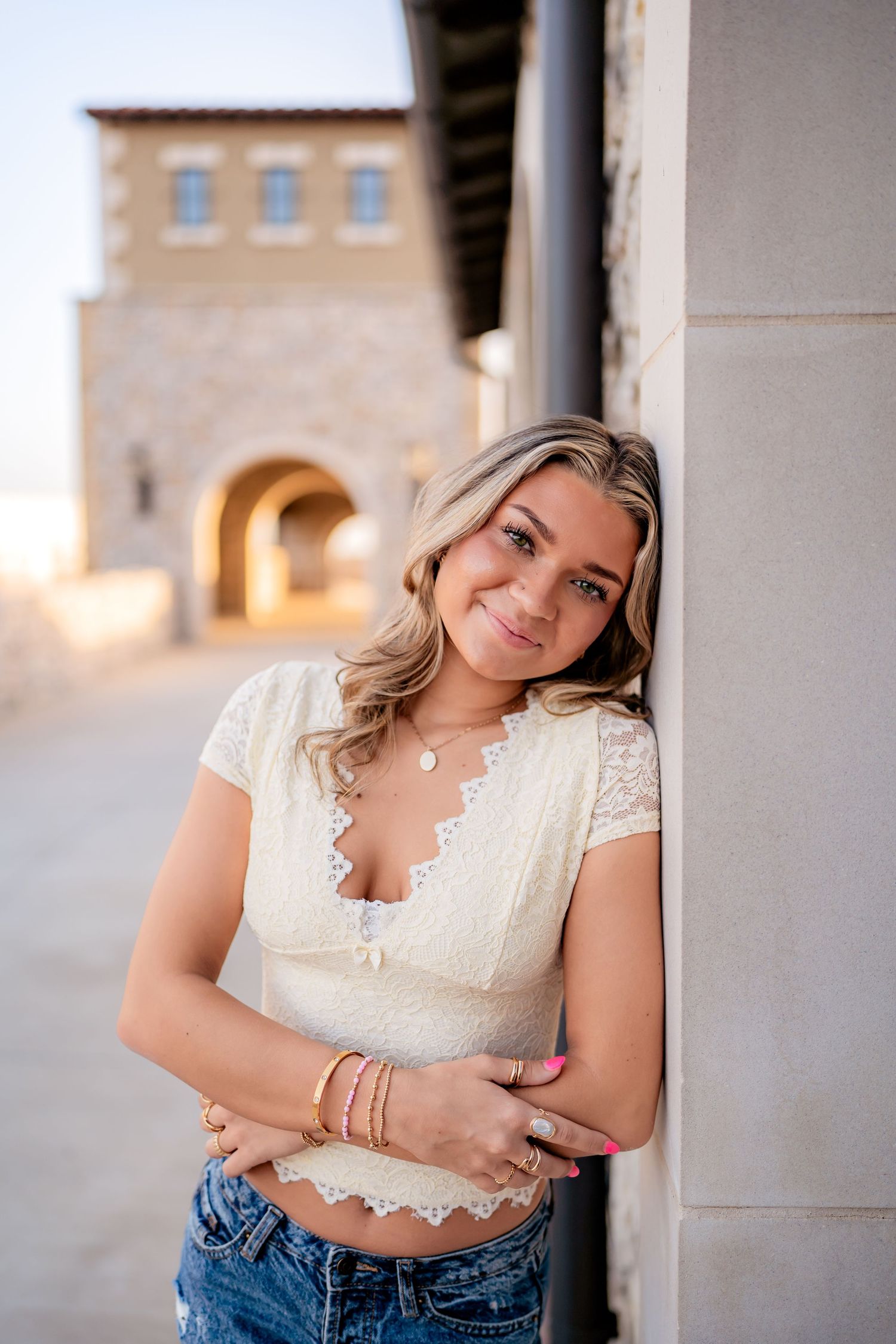 A young woman in a white lace crop top and jeans leans against a wall with Spanish-style architecture in the background.