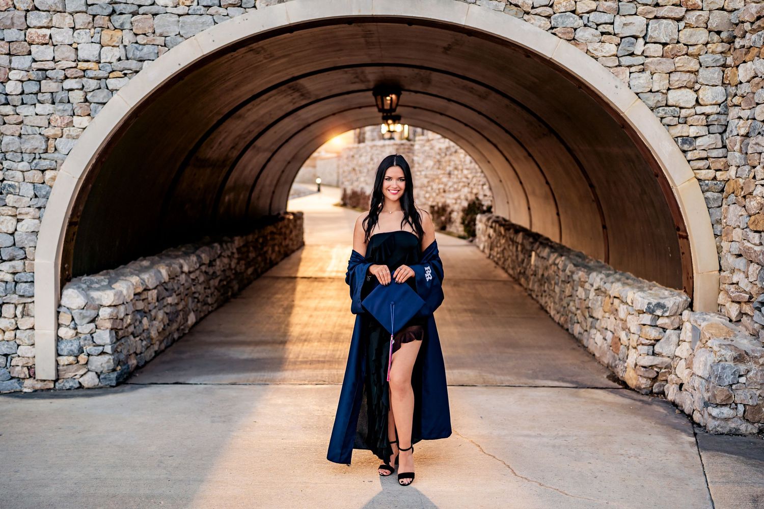 Person in an elegant navy blue dress poses in a stone tunnel with dramatic natural lighting shining through the archway.