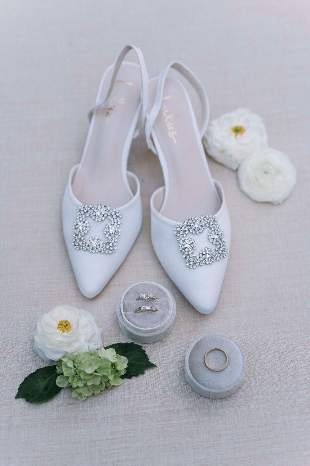 White bridal shoes with crystal embellishments displayed with wedding rings and delicate white and green flowers.