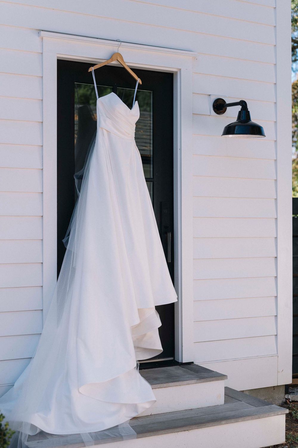 A white wedding dress hangs on display against a white clapboard wall with black light fixture.