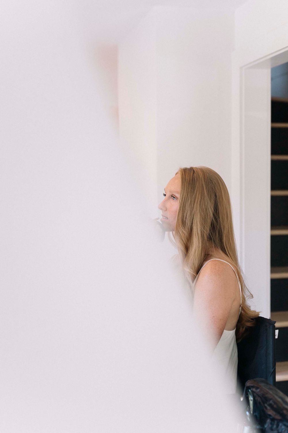 Person with long blonde hair leaning against a white wall in contemplative pose.