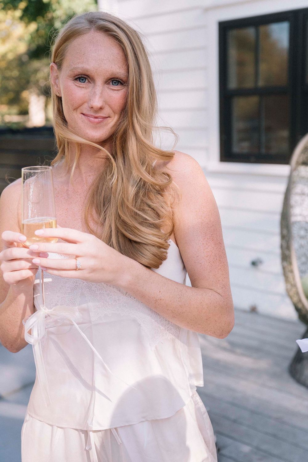 A woman in a light colored dress holds a champagne glass while standing outside.