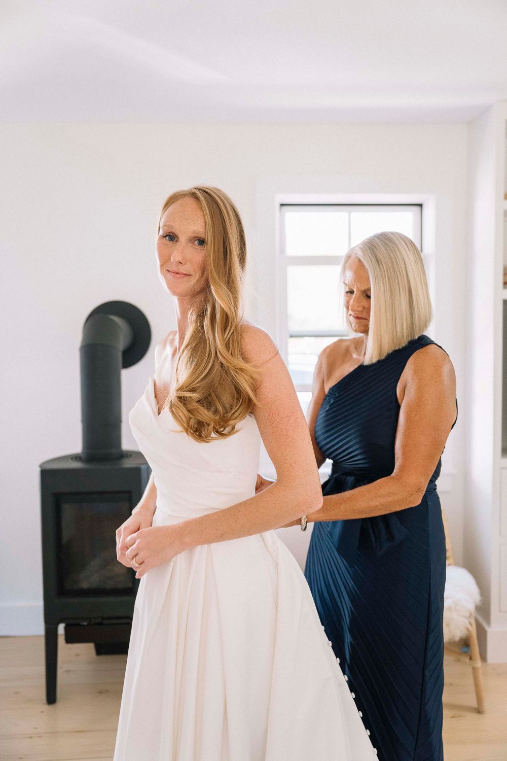 A pristine white wedding dress being adjusted in a room with a black wood stove.