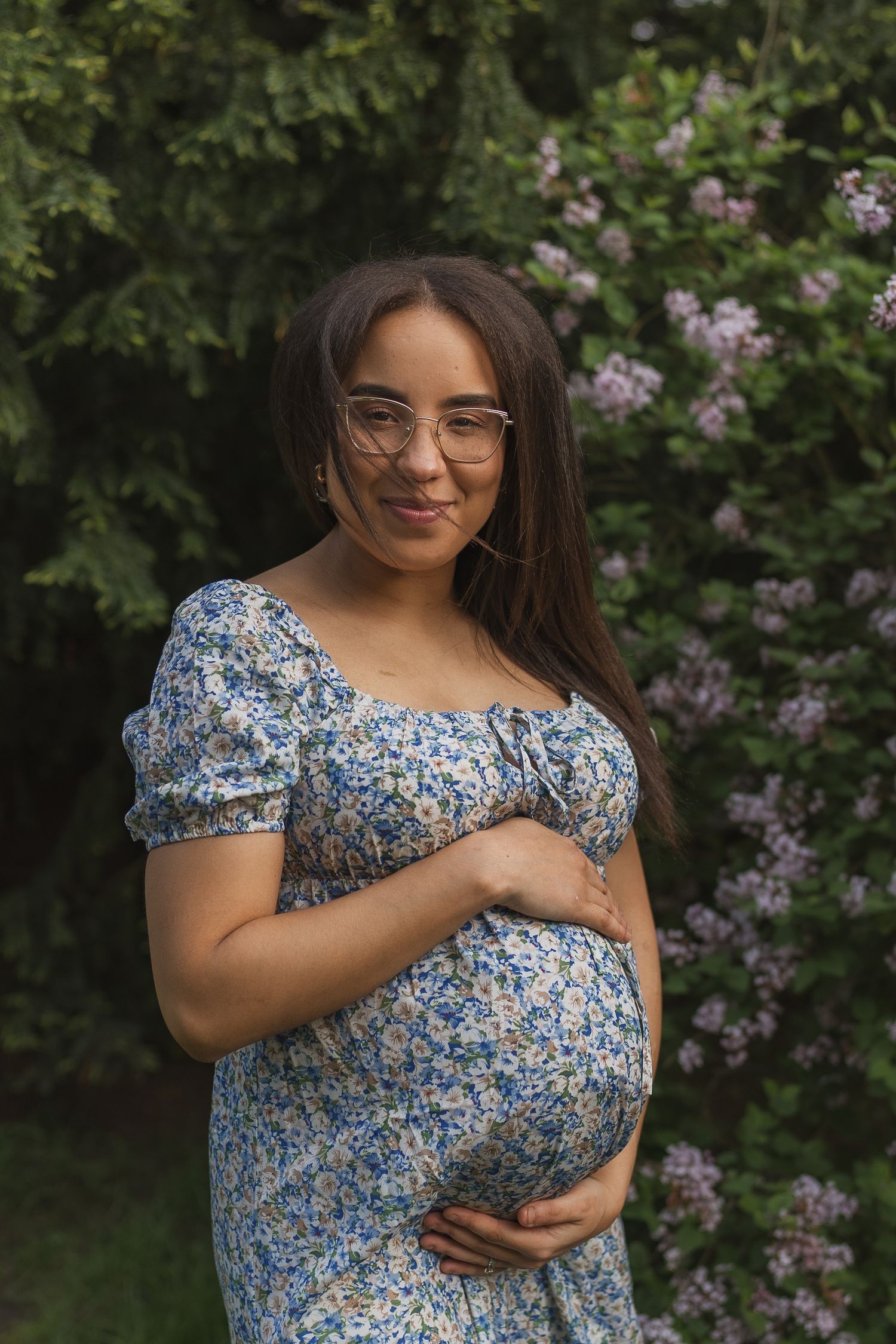 Expecting mother in a blue floral dress and glasses poses in a garden with blooming lilac flowers in the background.