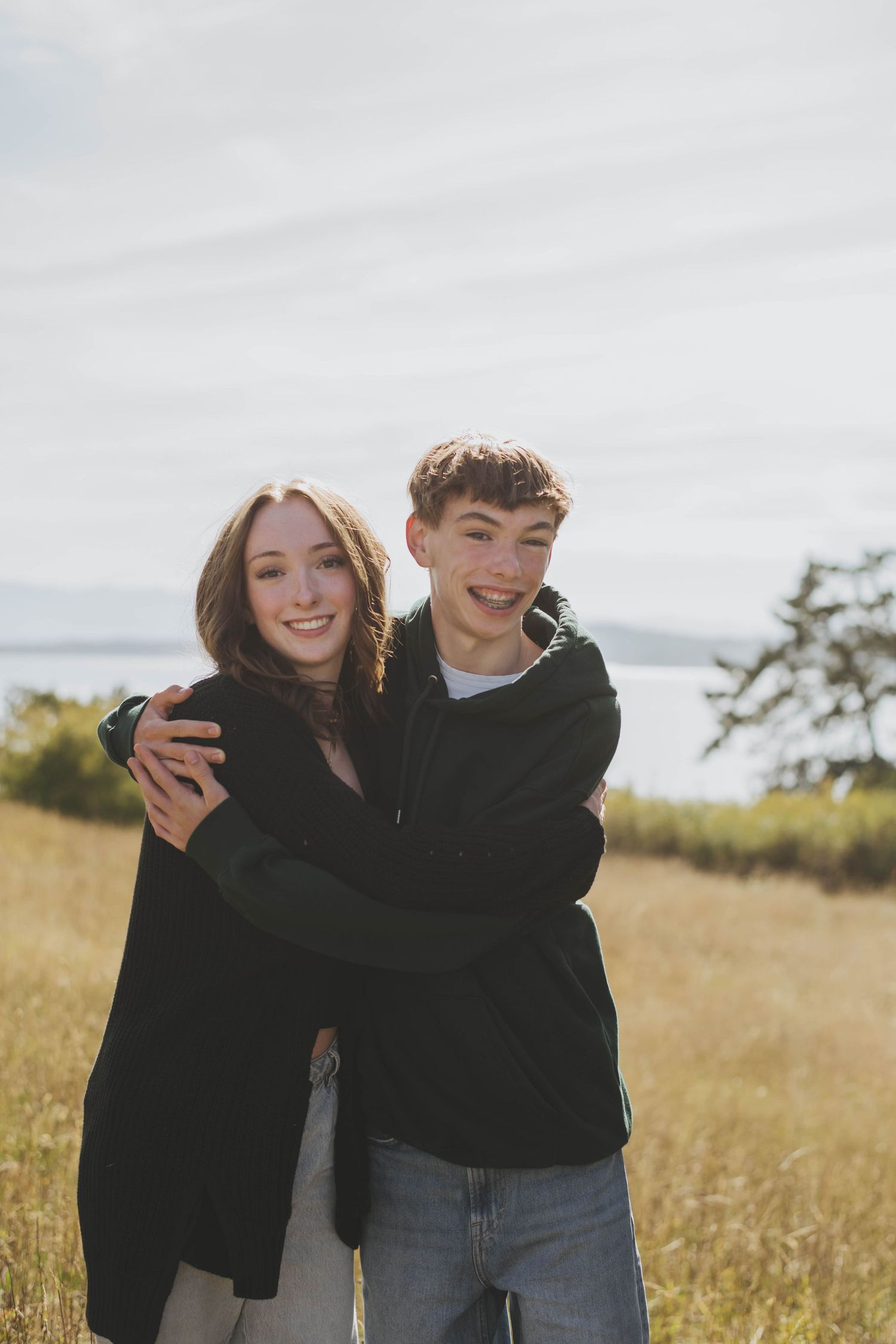 Twin siblings embrace and laugh together while standing in a golden field on an overcast day.