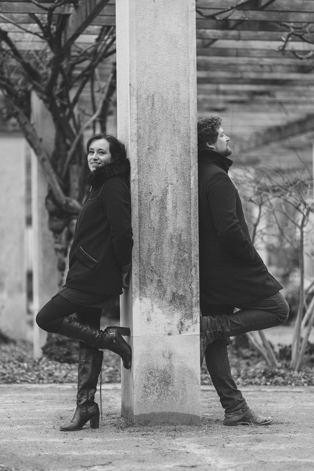 Black and white photo of two people posing back to back against a concrete pillar in an urban setting.
