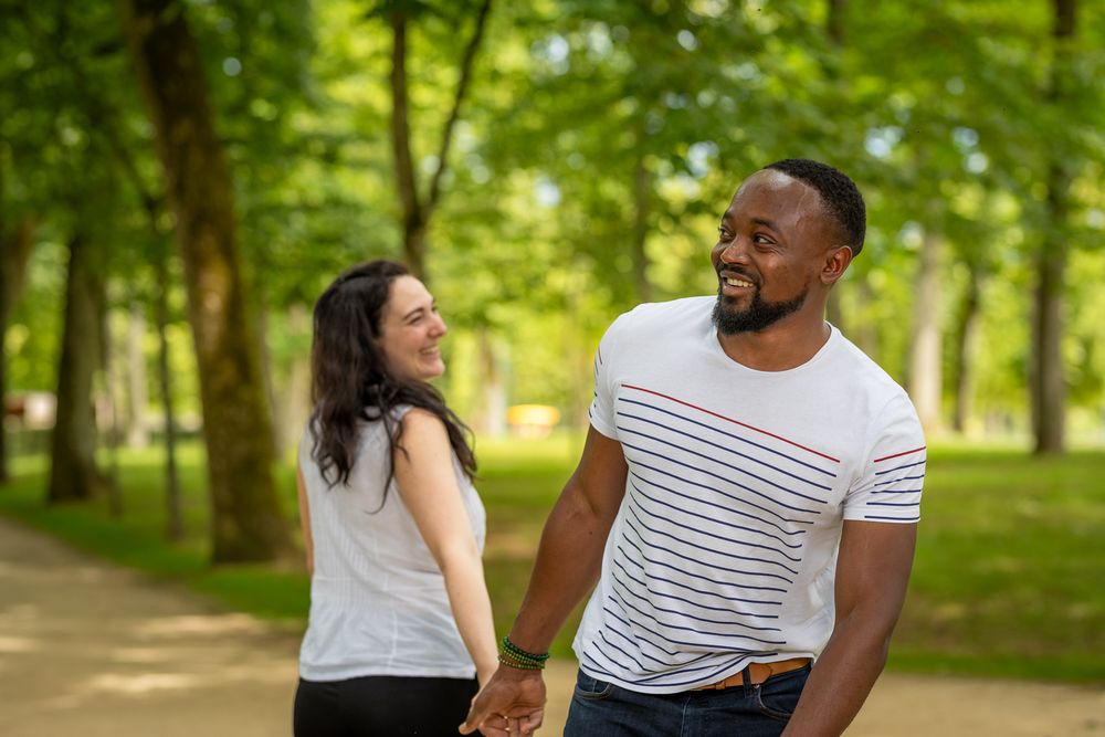 A happy couple walks hand in hand along a sunlit path in a lush green park on a summer day.