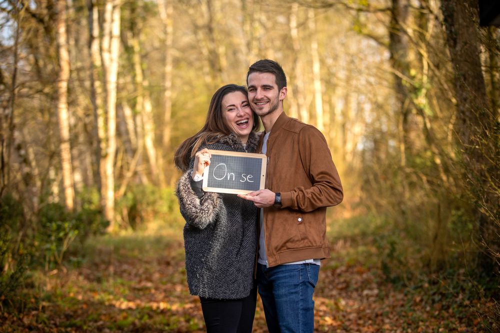 A couple stands in a wooded forest path holding a chalkboard sign on a sunny autumn day.