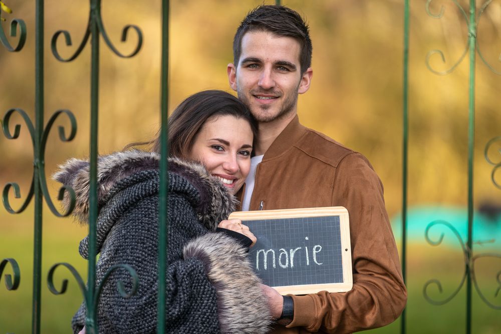 A couple posing outdoors holding a small chalkboard sign with a decorative iron gate in the background.