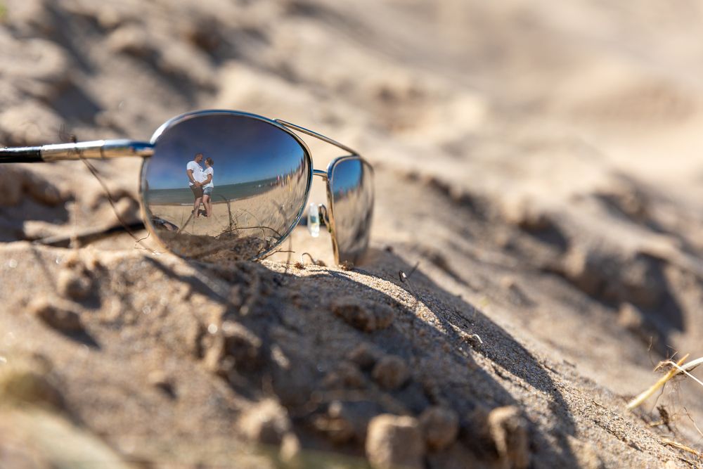 Pair of silver aviator sunglasses lying on sandy ground reflecting bright blue sky above.