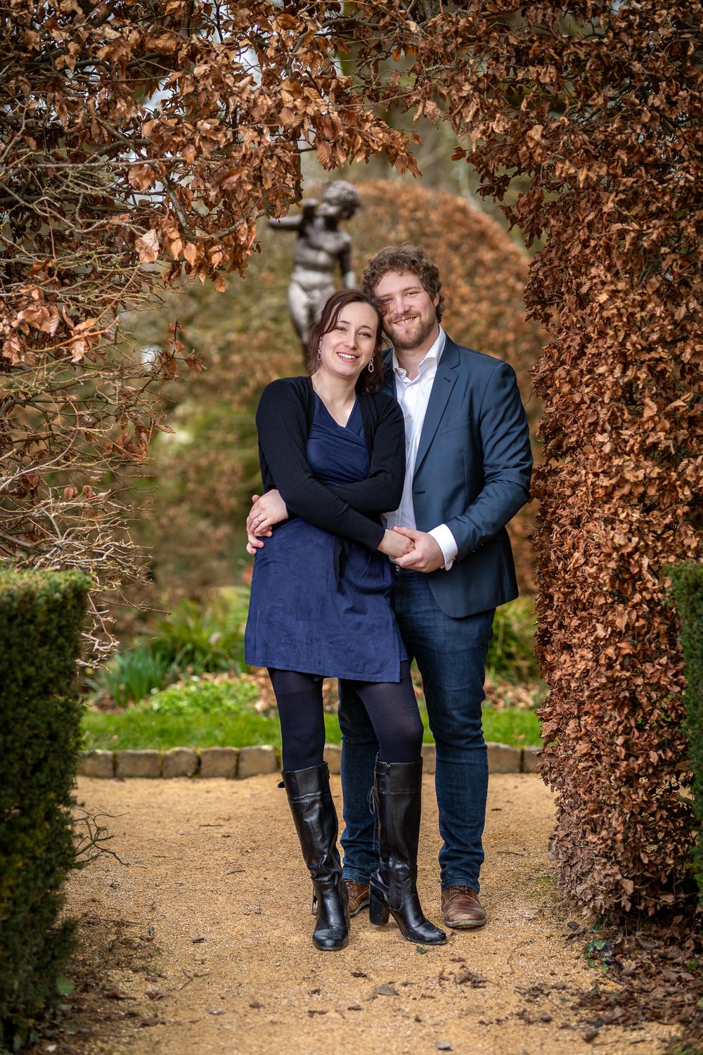 A couple poses together on a garden path framed by brown autumn foliage in a natural outdoor setting.