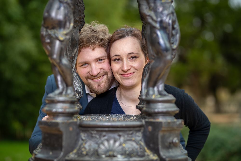 A couple peeks through an ornate metal garden sculpture with greenery in the background.