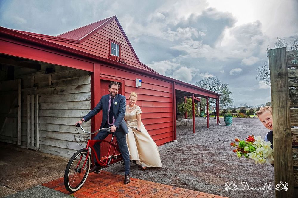 A couple poses with a vintage bicycle outside a red barn-style building on a cloudy day.
