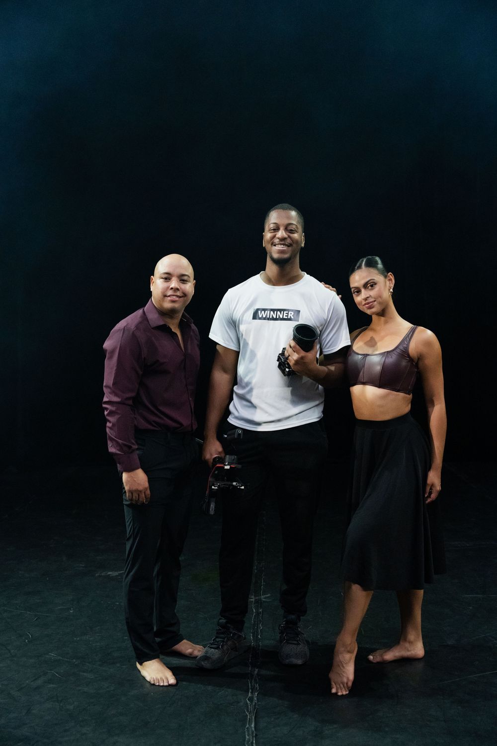 Three dancers in rehearsal attire pose together against a dark backdrop in a dance studio setting.