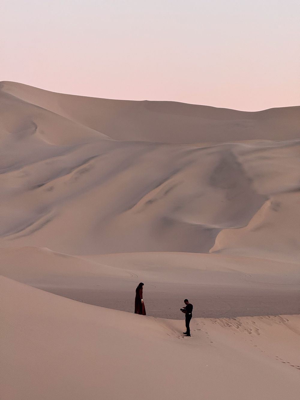 Two silhouetted figures stand on vast sandy dunes during a pink-hued sunset in the desert landscape.