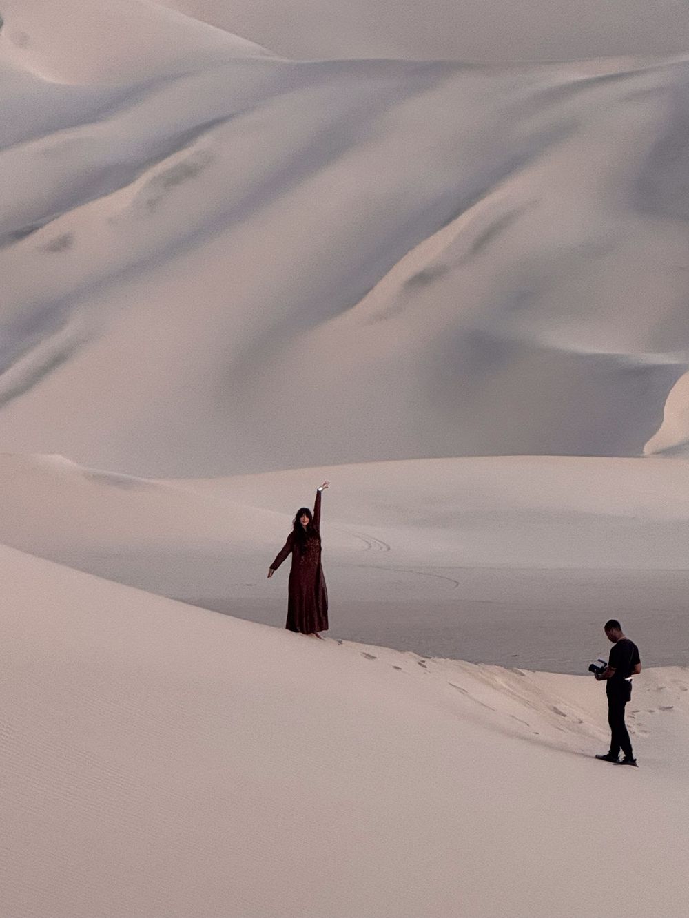 Two silhouetted figures stand among smooth, snow-covered sand dunes creating a dramatic winter desert landscape.