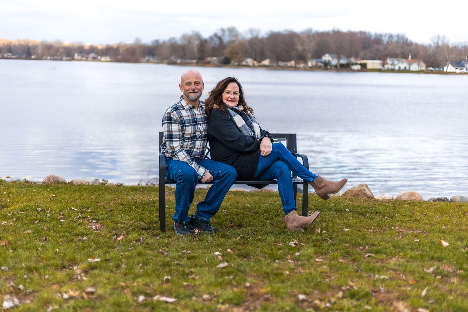 Two people sit together on a blue bench by a serene lake on a fall day with autumn trees in the background.