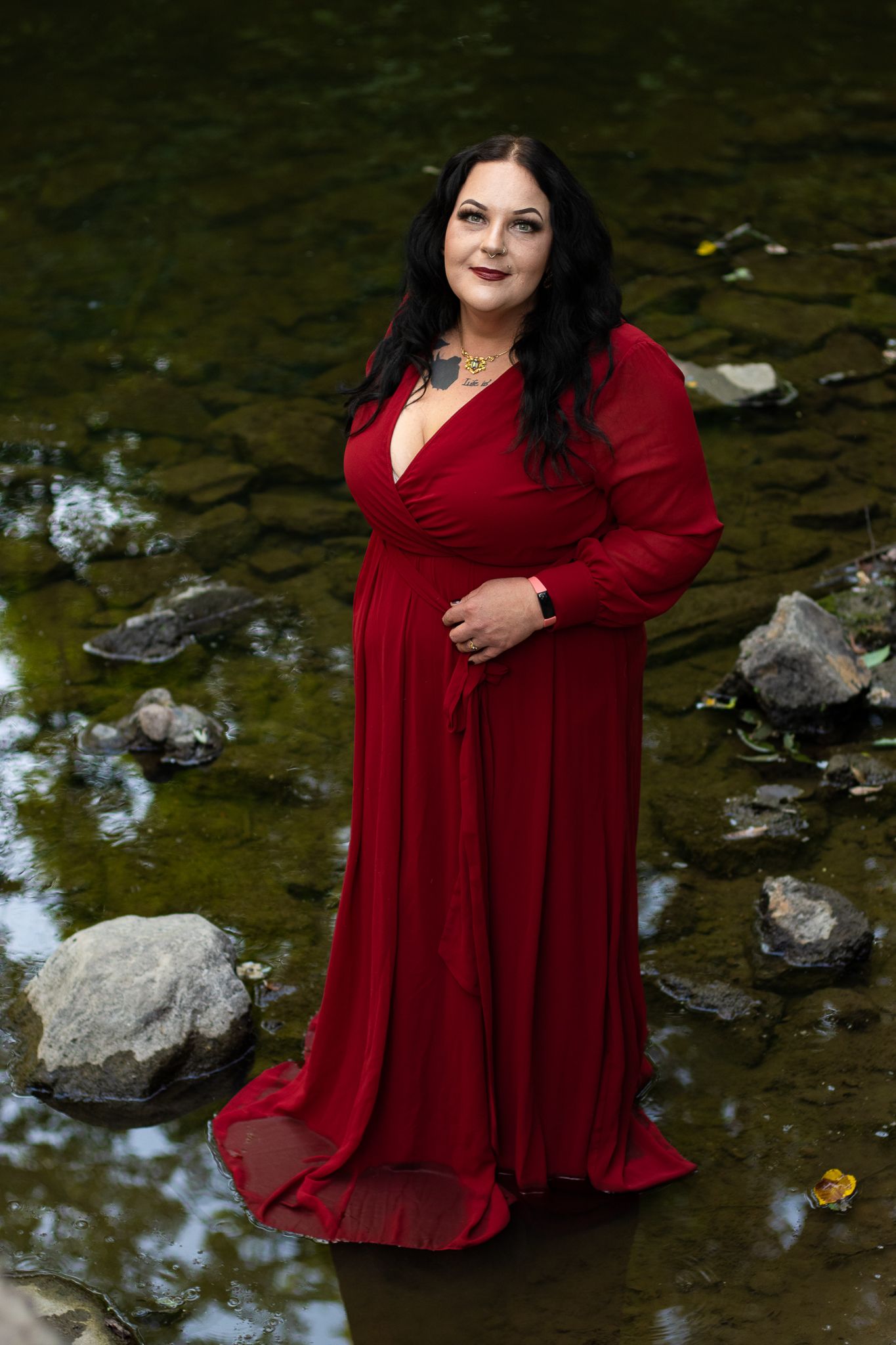 A flowing red maxi dress is worn in a nature setting with rocks and a stream, creating an elegant outdoor portrait.