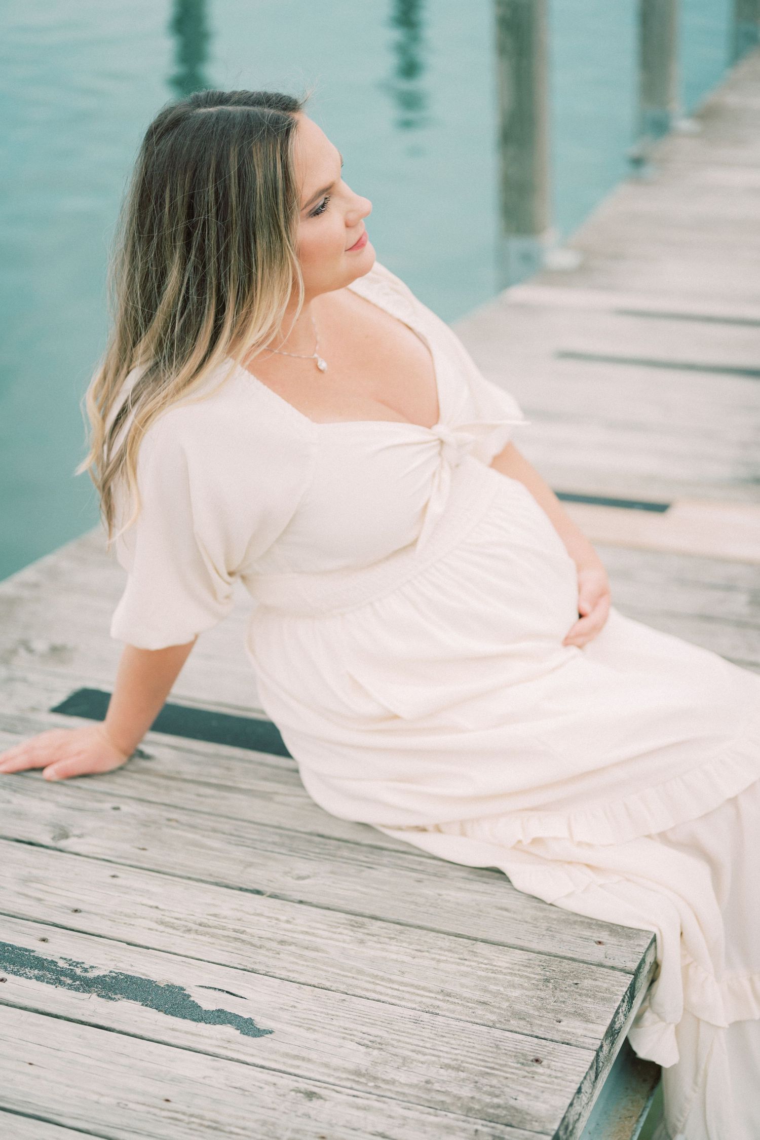 Pregnant woman in white dress poses serenely on wooden dock by waterfront.